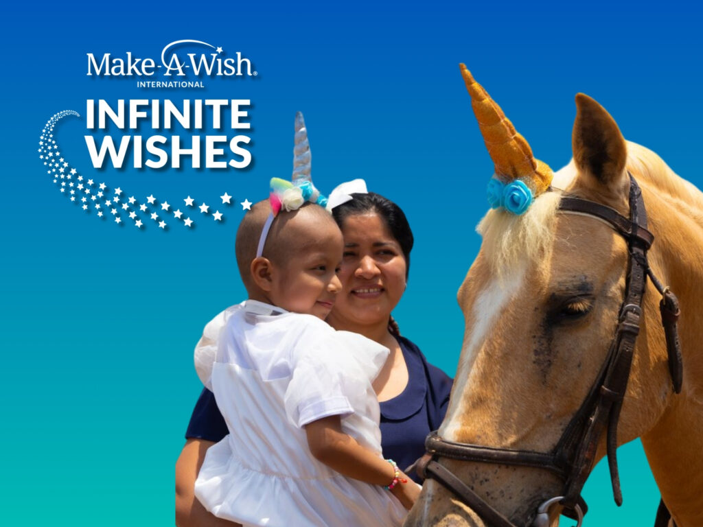 A smiling woman holds a young child wearing a unicorn headband, next to a horse with a unicorn horn against a blue background with the Make-A-Wish International Infinite Wishes logo.