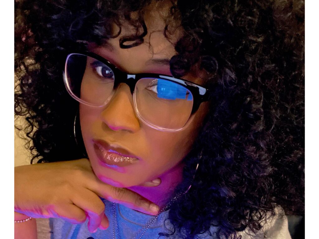 A person with curly hair and large glasses rests their chin on their hand, looking at the camera. Light reflects off their glasses, highlighting their glossy lips and hoop earrings.