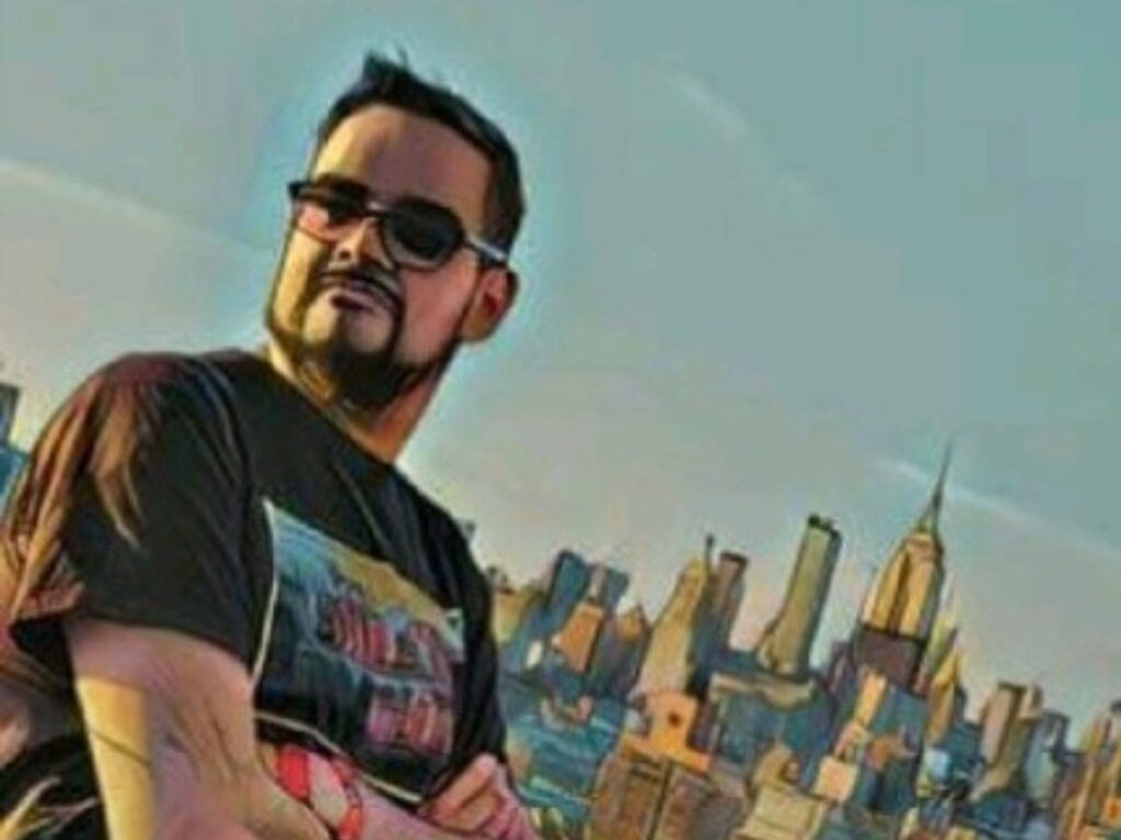 A man with short dark hair, a beard, and sunglasses stands with his arms crossed in front of a city skyline under a clear sky. The image appears to have a digital, painting-like filter.
