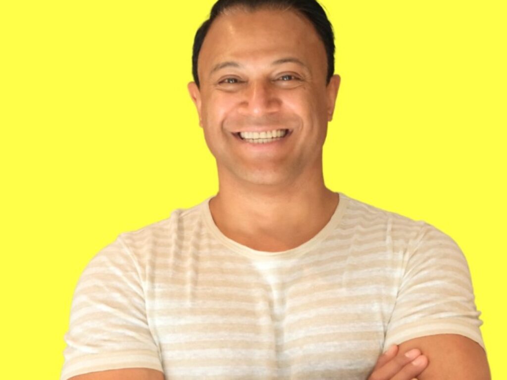 A smiling person with short dark hair, wearing a light-colored striped t-shirt, stands with arms crossed against a bright yellow background.