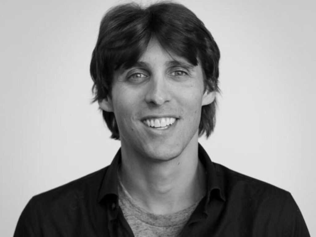 A man with medium-length dark hair smiles at the camera. He is wearing a dark collared shirt over a light t-shirt, against a plain light background. The image is in black and white.