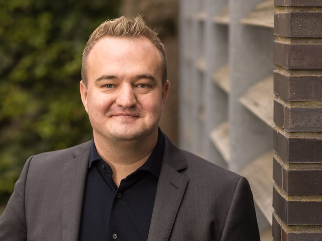 A man with short, light brown hair is wearing a dark suit and black shirt, smiling while standing outdoors near a brick wall and greenery.