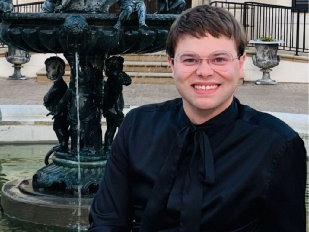 A person with short brown hair, glasses, and a black shirt smiles while sitting in front of a decorative fountain outdoors. The background includes steps, railings, and large planters.