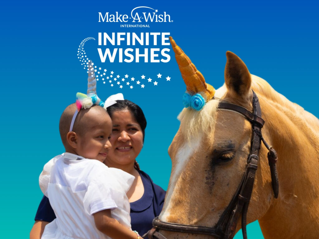 A smiling child wearing a unicorn headband and a white dress sits with an adult beside a horse also wearing a unicorn horn, beneath a Make-A-Wish International: Infinite Wishes logo on a blue gradient background.