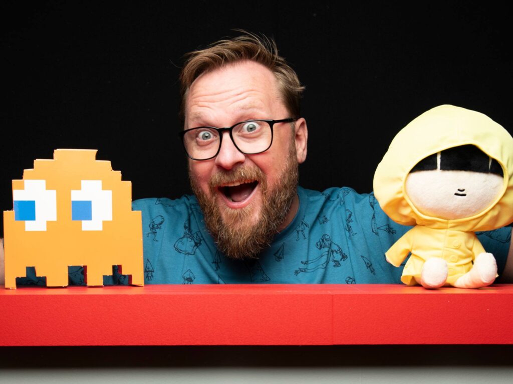 A bearded man with glasses and a blue shirt smiles excitedly behind a red surface, between a pixelated orange ghost figure and a plush toy in a yellow hooded outfit. The background is black.