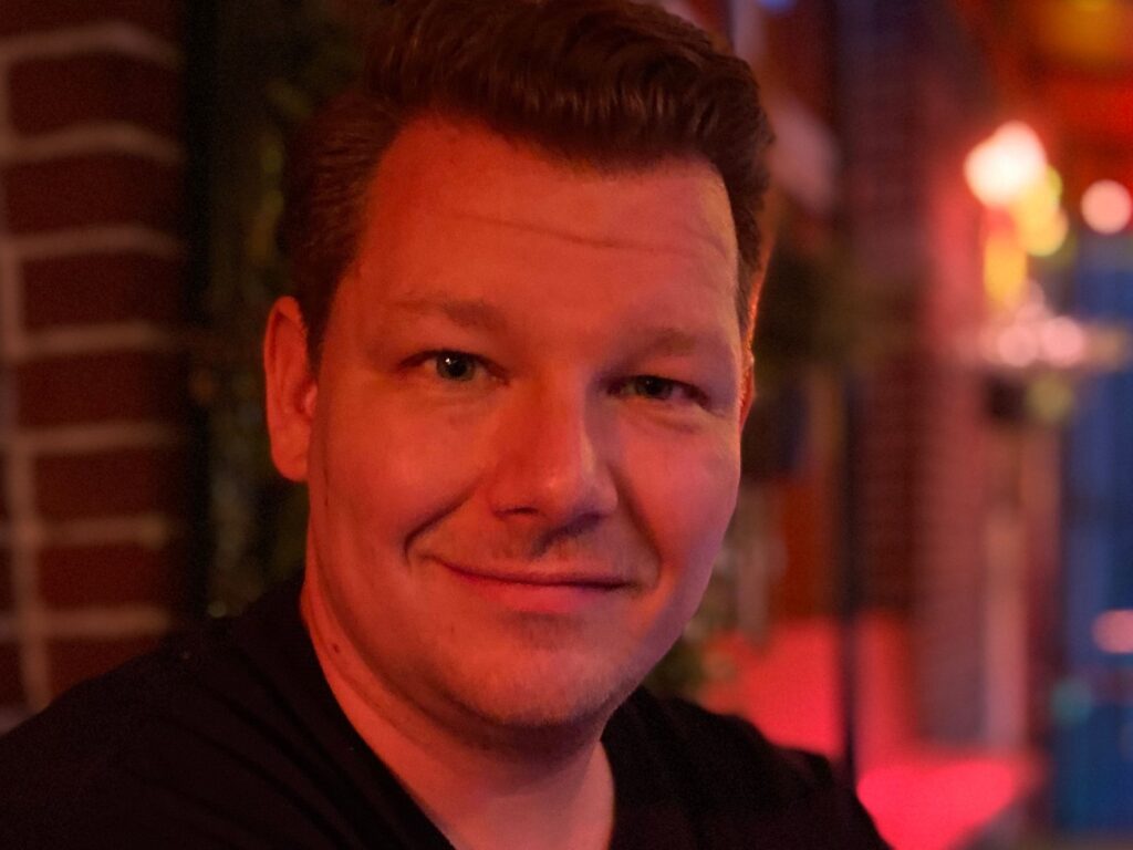 A man with short, light brown hair and a slight smile is sitting indoors, with warm red and orange lighting and blurred background elements, including brick walls and plants.