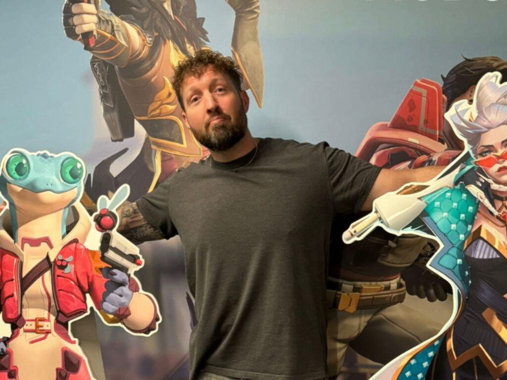 A man with curly hair and a beard stands with his arms outstretched, posing between two large cartoon character cutouts—a blue lizard in a red jacket and a white-haired character in colorful armor.