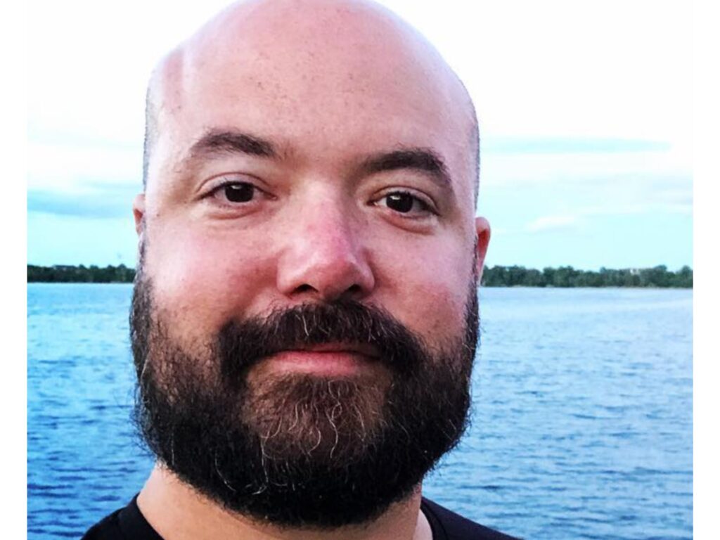 A bearded man with a shaved head is standing outdoors near a body of water, with trees and a blue sky in the background.
