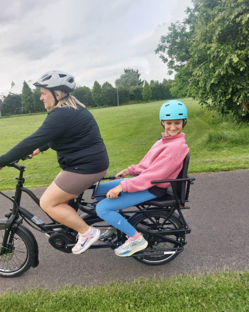 Two people ride a bike on a paved path through a park. The adult in front wears a helmet and black jacket, while the smiling child at the back wears a blue helmet and pink sweater, sitting comfortably. Green grass and trees are visible.