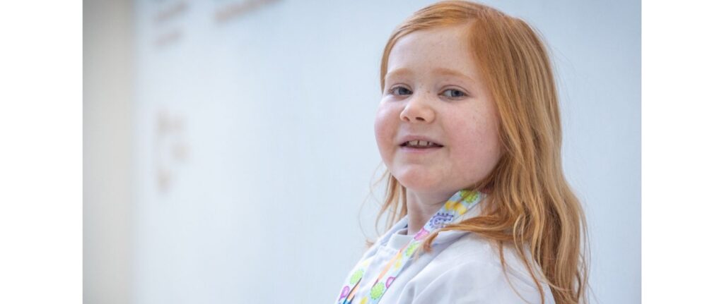 A young girl with long red hair smiles softly at the camera, wearing a white shirt and a colorful lanyard, against a plain light background.