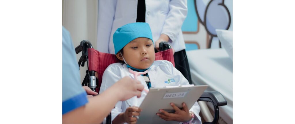 A young child in a wheelchair, dressed in a blue surgical cap and white coat, holds a clipboard labeled BED 23 while adults in medical attire stand nearby in a hospital setting.