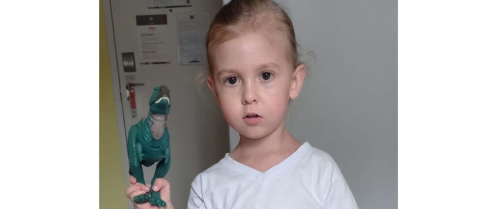 A young child with light brown hair, wearing a white shirt, stands indoors holding a green toy dinosaur and looking directly at the camera with a neutral expression.