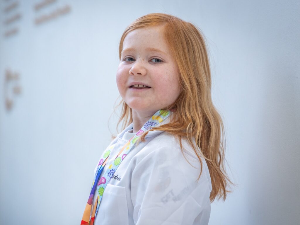 A young girl with long red hair, wearing a white lab coat and a colorful lanyard, smiles softly while standing against a light-colored wall, reflecting Audrinas wish to be a scientist.