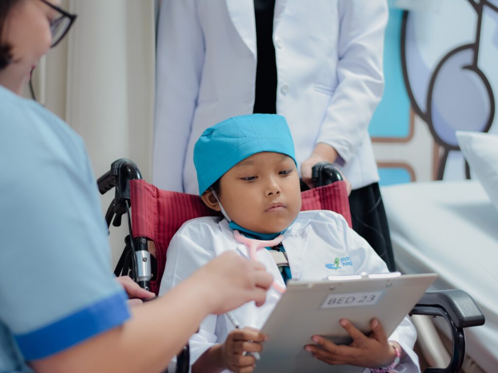 A child in a wheelchair, dressed as a doctor with a stethoscope and blue cap, holds a clipboard labeled BED 23 while an adult in scrubs points at it. Another adult in a white coat stands nearby.