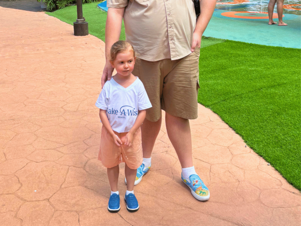 A young girl wearing a Make-A-Wish shirt and peach shorts stands beside an adult in a tan outfit on a paved walkway near artificial grass, capturing the heartwarming spirit of João Arturs wish to go to a dinosaur park. The adult’s hand rests gently on her shoulder.