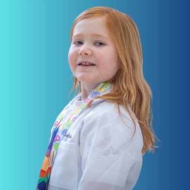 Young girl with long red hair, wearing a white lab coat and a colorful scarf, smiles softly while standing in front of a blue gradient background.
