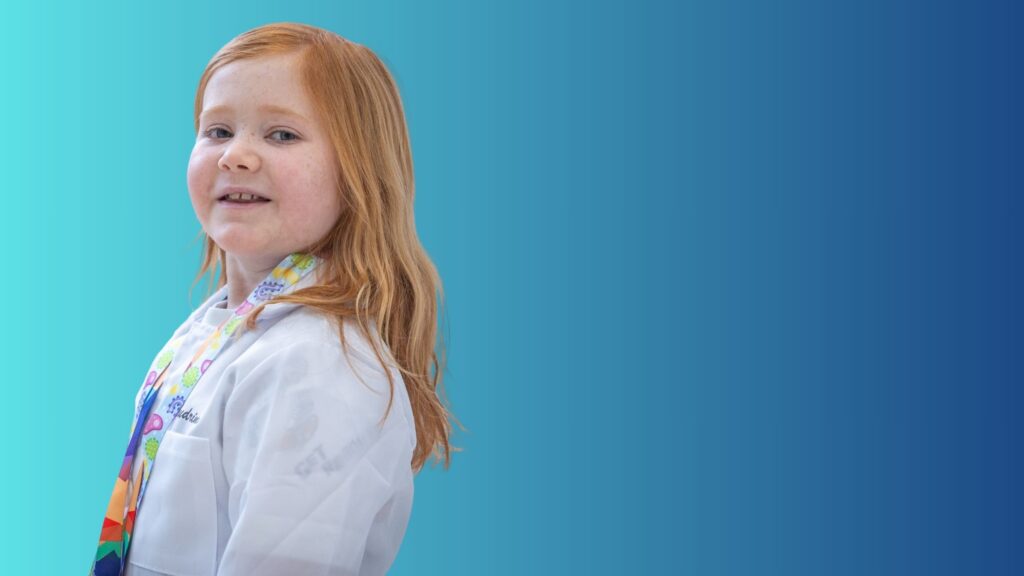 A young girl with long red hair smiles slightly and looks toward the camera. She is wearing a white lab coat and a colorful patterned scarf, standing against a blue gradient background.