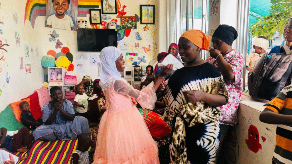 A group of children and adults gather in a colorful room decorated with wall art and drawings. Two women in the center greet each other warmly, while others watch and interact around them.