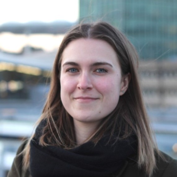 A young woman with straight brown hair and blue eyes, wearing a dark scarf and coat, stands outdoors with a blurred cityscape and tall buildings in the background. She is smiling softly at the camera.