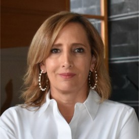 A woman with shoulder-length blonde hair, wearing a white blouse and large hoop earrings, stands indoors with a neutral expression. The background includes a dark wall and wooden elements.