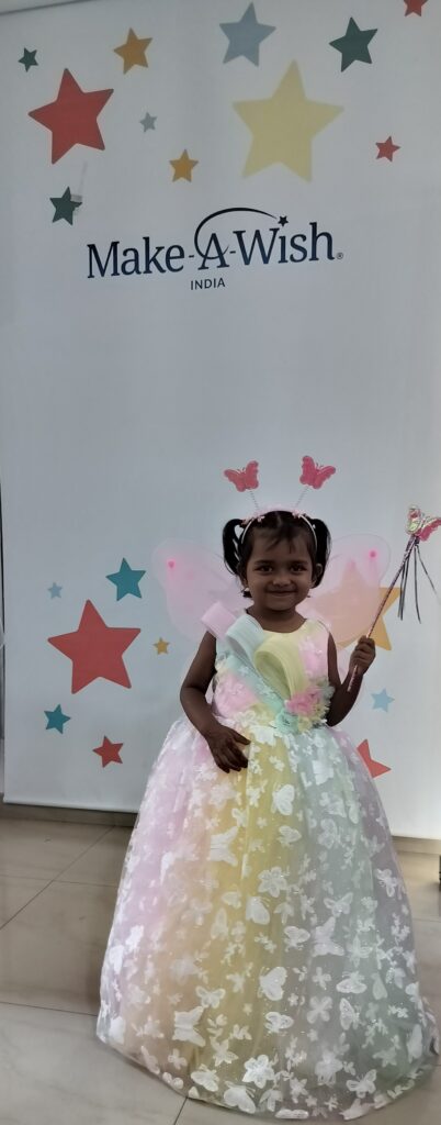 A young girl, granted Mantasha's wish to have a fairy dress, smiles in a colorful gown and butterfly headband while holding a wand before a Make-A-Wish India backdrop adorned with colorful stars.