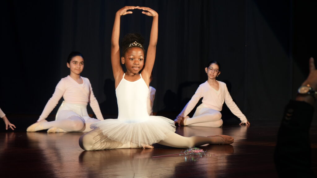A young girl in a white tutu performs a split with arms raised on stage, while other ballet dancers in pale costumes sit behind her, smiling. The stage is lit and the background is dark.