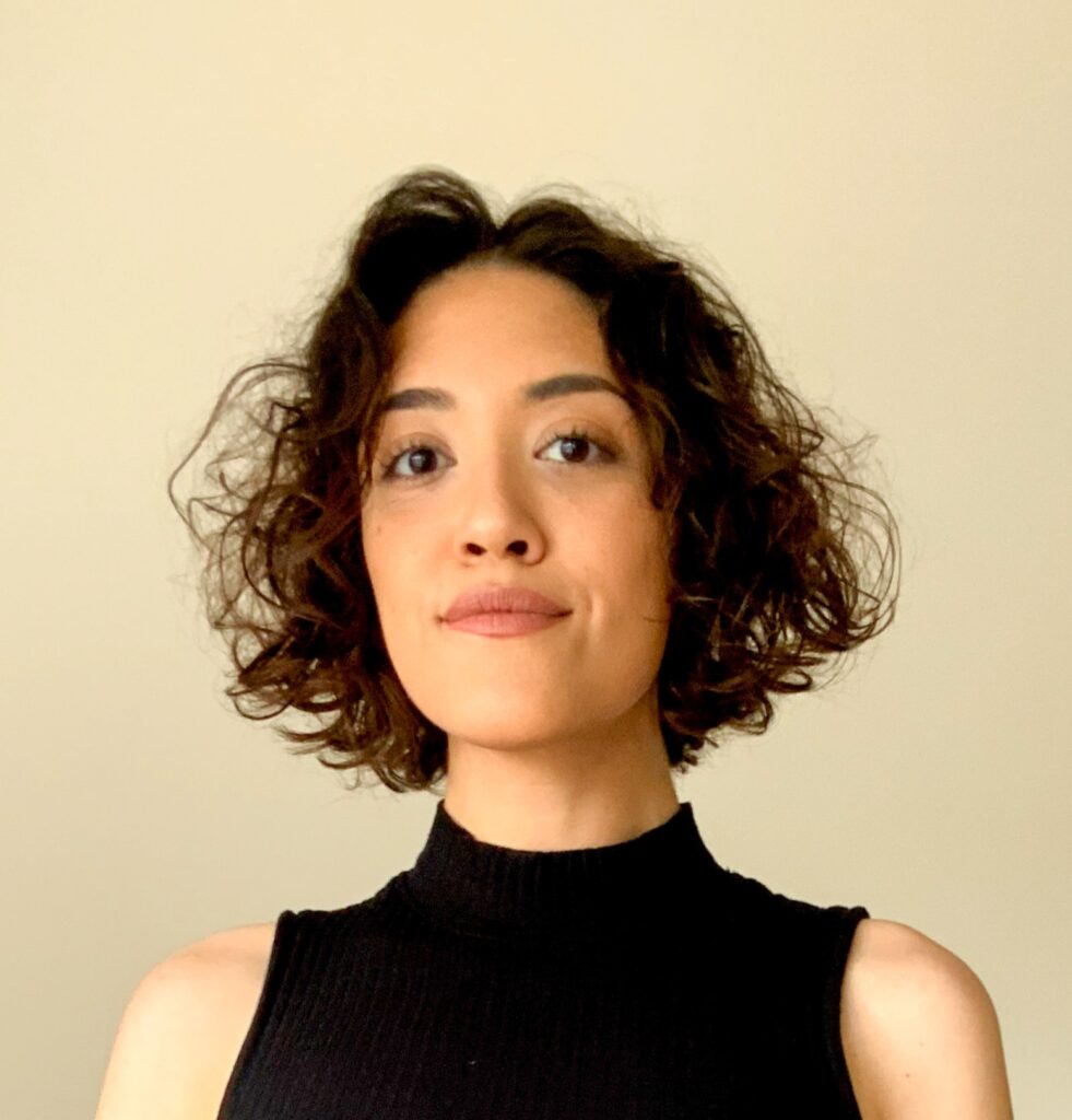 A person with short, curly brown hair wearing a sleeveless black turtleneck top, standing in front of a plain beige background.
