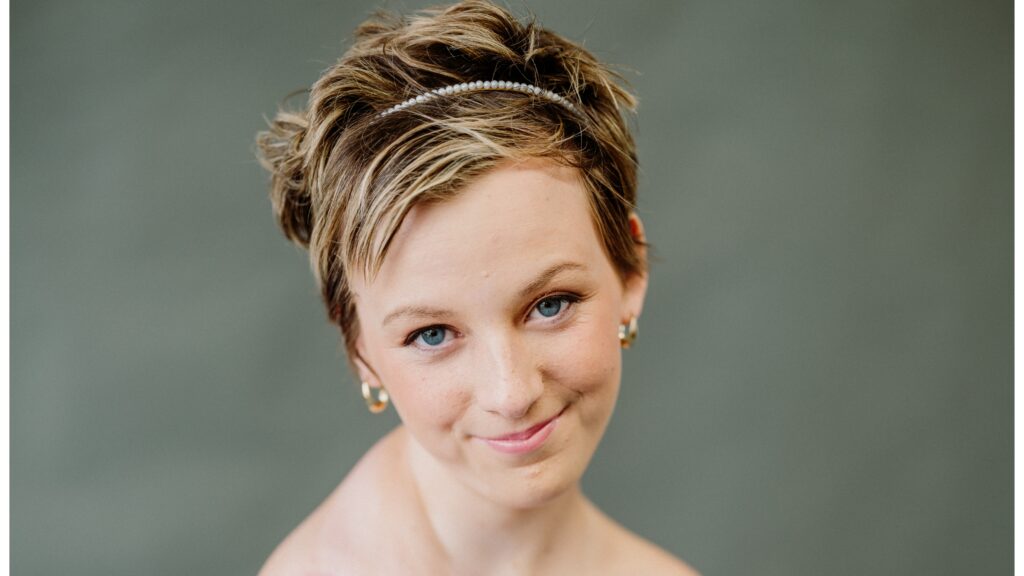 A person with short, light brown hair wears a pearl hairband and gold earrings, smiling softly at the camera. The background is solid and muted green, capturing the gentle anticipation of Rylee’s wish to have a shopping spree.