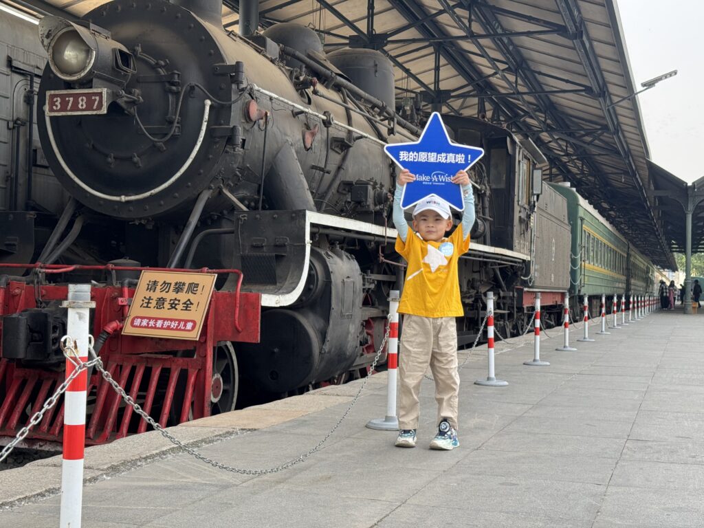 A young child in a yellow shirt and blue hat stands on a train platform, holding a blue star-shaped sign, with a vintage steam locomotive and passenger cars in the background.