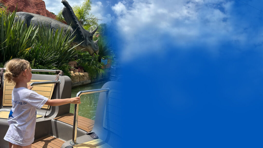 A young girl stands on a boat ride at a theme park, looking at a large dinosaur model surrounded by greenery; the right side of the image fades into a solid blue area and sky with clouds.