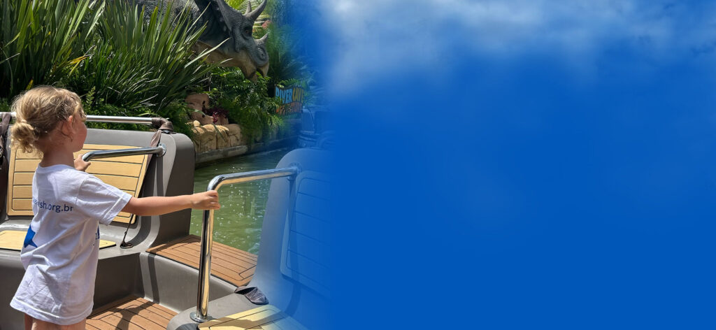 A young child with curly blond hair, wearing a white t-shirt, stands on a boat ride in a theme park, looking toward greenery and a dinosaur statue, with a large blue area overlaying the right side of the image.