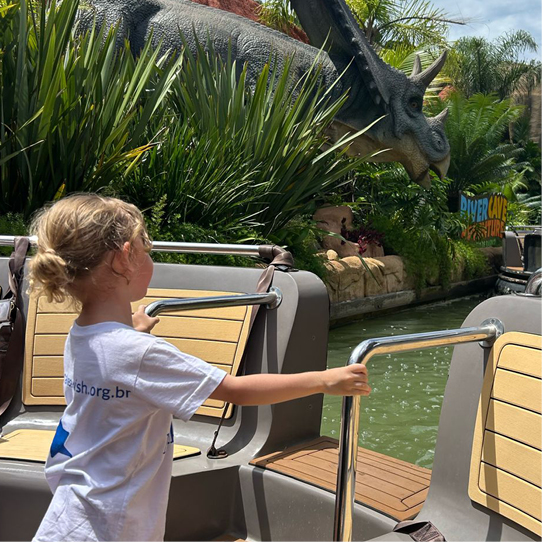 A young child on a boat ride looks at a large triceratops dinosaur statue surrounded by greenery at a theme park. The sky is clear and sunny.