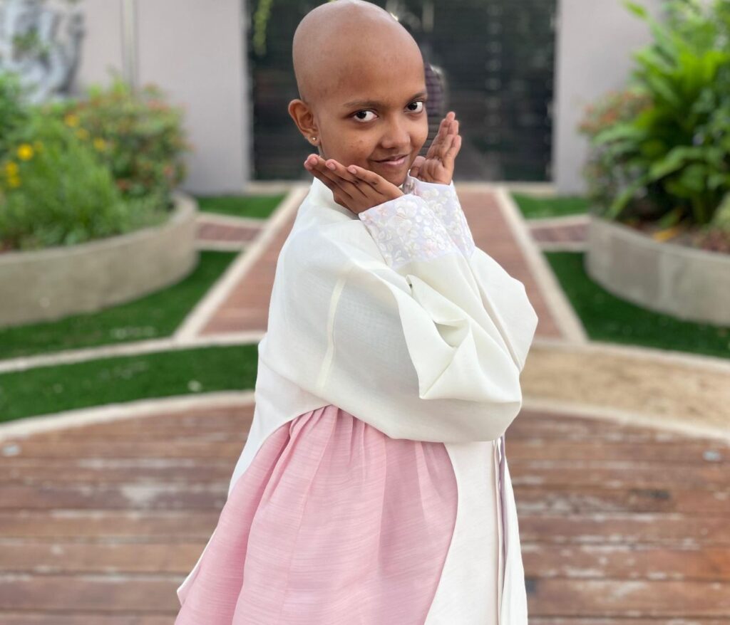 A young girl with a bald head poses playfully outdoors, wearing a light pink dress with a white overcoat. She stands on a wooden deck, with greenery and a walkway in the background.
