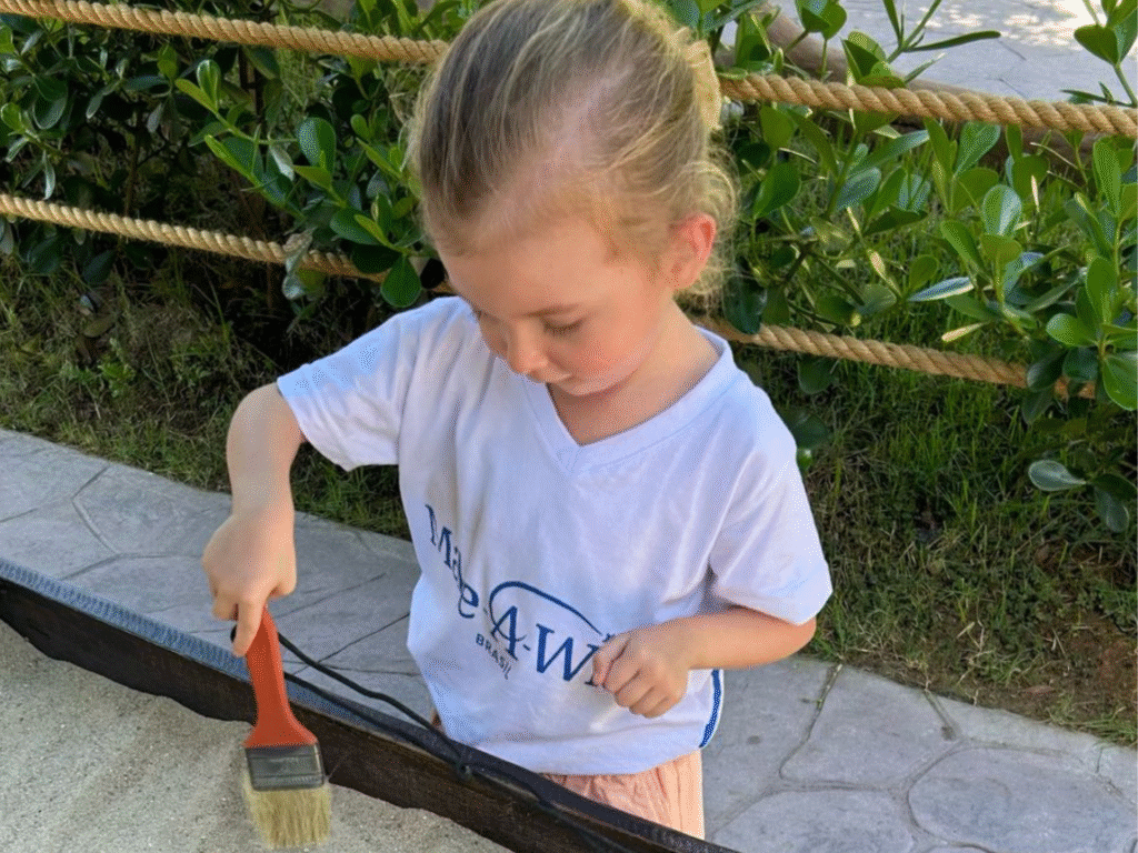 A young child with blonde hair wearing a white t-shirt is using a paintbrush to brush sand inside a bordered area outdoors, with green bushes and rope fencing in the background.