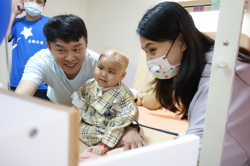 A young child with a bandage on their head sits on a hospital bed, flanked by two smiling adults. The adults wear masks, and the child has medical equipment attached. Another person stands in the background.
