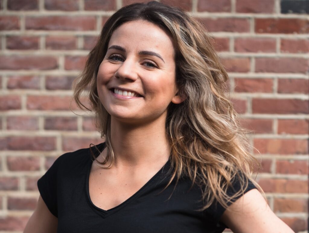 A smiling woman with wavy hair stands in front of a brick wall, wearing a black t-shirt. She has her hands on her hips and looks confident.