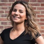 A smiling woman with wavy hair stands in front of a brick wall, wearing a black t-shirt. She has her hands on her hips and looks confident.