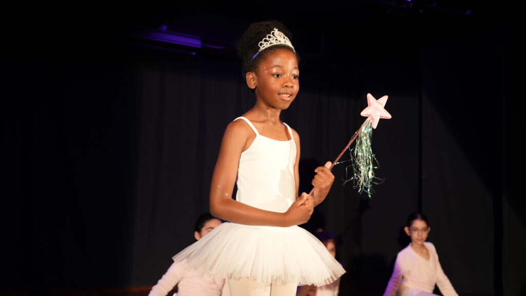 A young girl dressed as a ballerina fairy, wearing a white tutu, a tiara, and holding a star wand, stands on stage with other children in the background.
