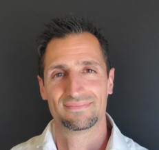 A man with short dark hair and a trimmed goatee, wearing a light-colored shirt, smiles softly in front of a plain dark background.
