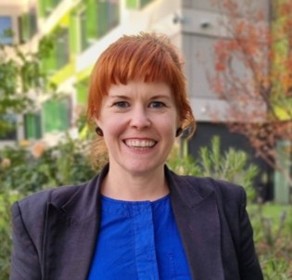 A smiling woman with short red hair, wearing a blue top and dark blazer, stands outdoors in front of greenery and a modern building with green and white accents.