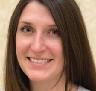 A woman with straight, long brown hair smiles at the camera. She has light skin, blue eyes, and is wearing a light-colored top. The background is plain and beige.