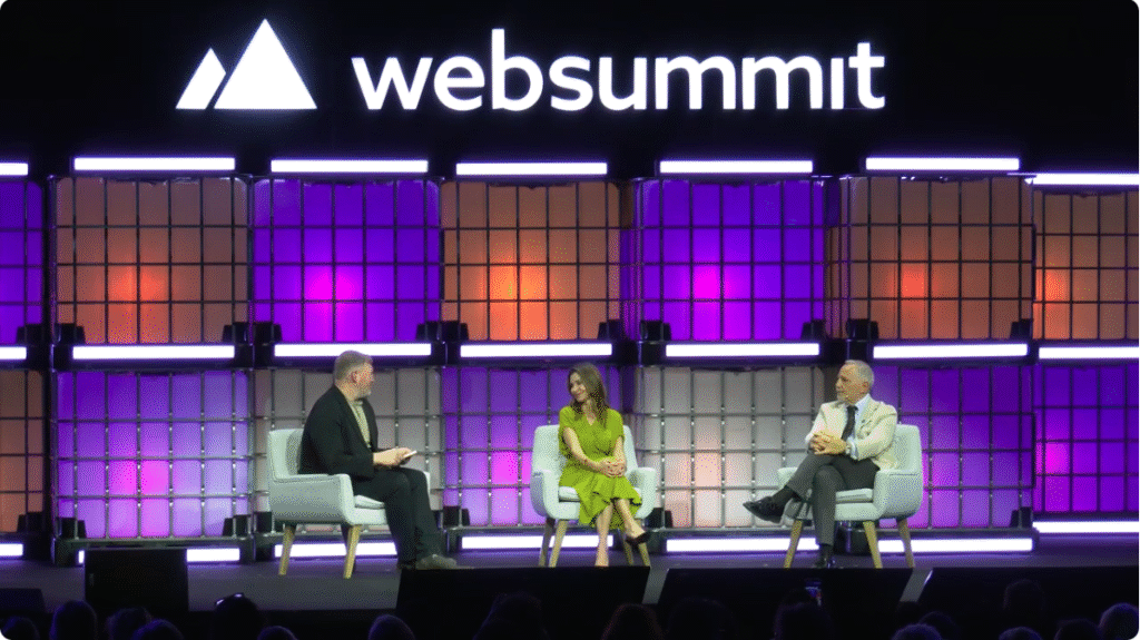 Three people sit on stage in chairs having a discussion at Web Summit. Bright purple and orange lights illuminate the backdrop, and the Web Summit logo is displayed above them.