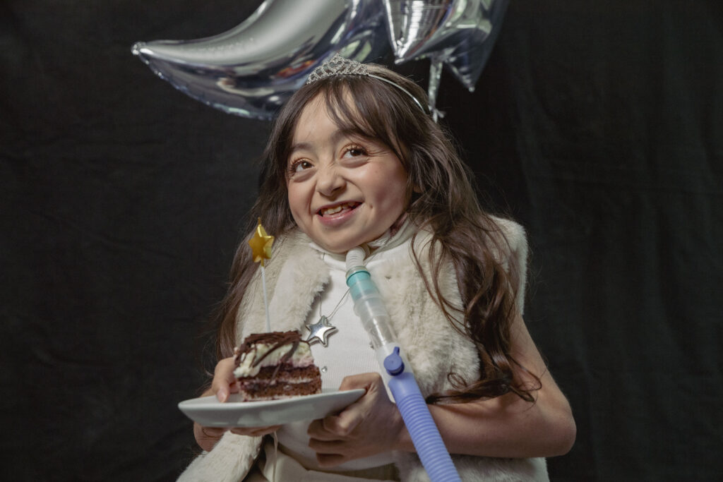 A young girl, part of Make-A-Wish International Appeals for Help to Unwrap the Power of a Wish This Holiday Season, smiles in a tiara and fur vest, holding chocolate cake and a star wand before silver balloons and a black background.