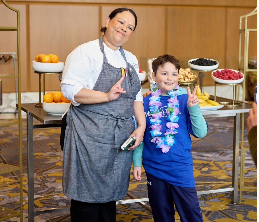 A chef in a gray apron and a child wearing a blue shirt and purple flower lei smile and make peace signs while posing together in front of a table with bowls of fruit at the Fairmont.