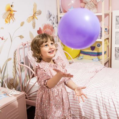 A young child in a pink dress smiles while tossing a purple balloon in a decorated bedroom with floral wallpaper and a striped bedspread.