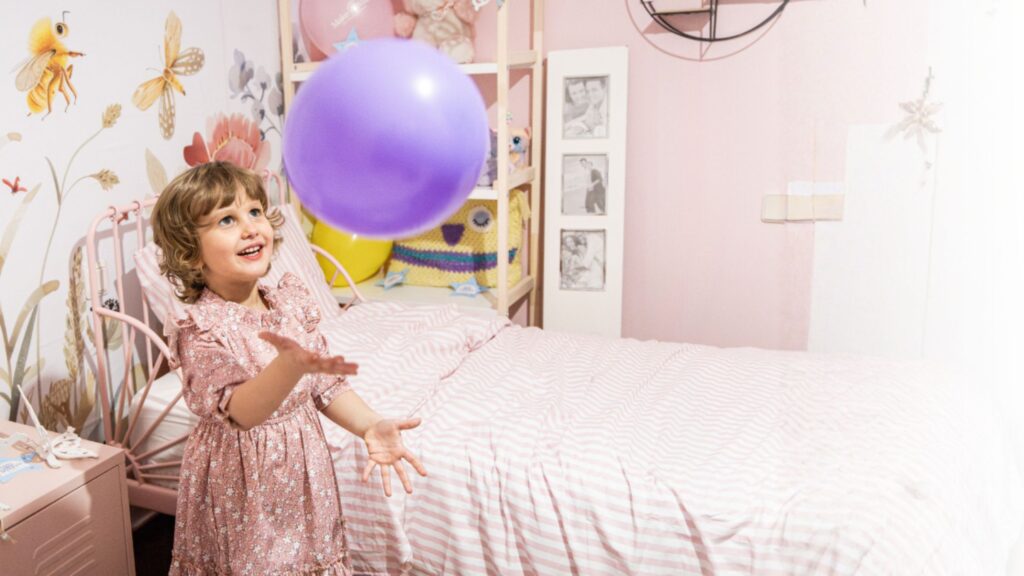 A young child in a pink dress smiles and tosses a purple balloon in a pastel-themed bedroom with butterfly wall decor and a neatly made bed.
