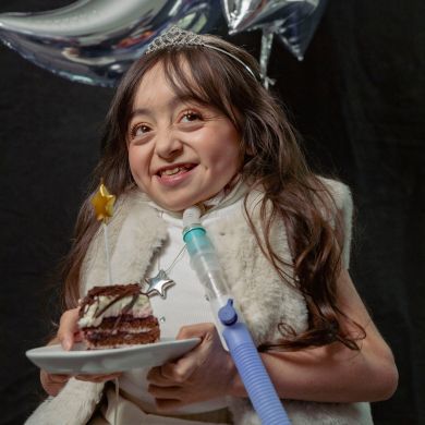 A young girl wearing a tiara and fur vest smiles while holding a plate with a slice of cake and a star-shaped candle. She has a medical breathing tube and is in front of silver balloon decorations.