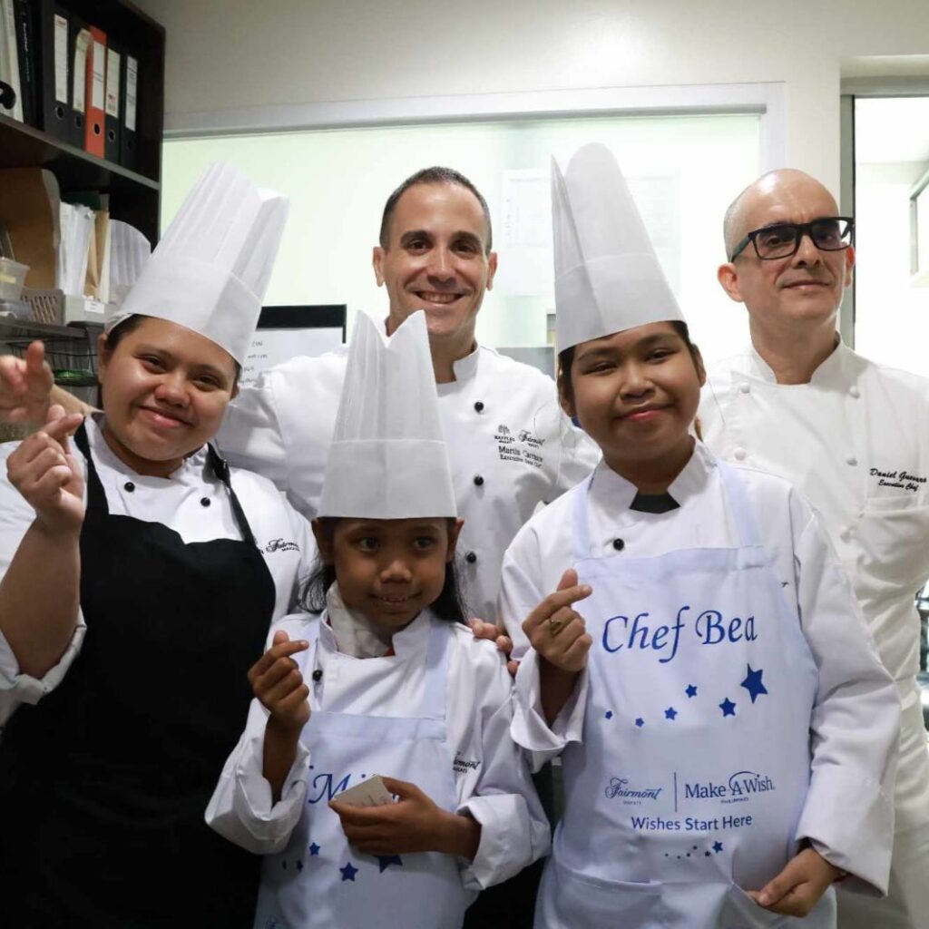Two chefs and three children in chef hats and aprons pose and smile together in a Fairmont kitchen; the children's aprons feature the Chef Bea and Make-A-Wish logos.
