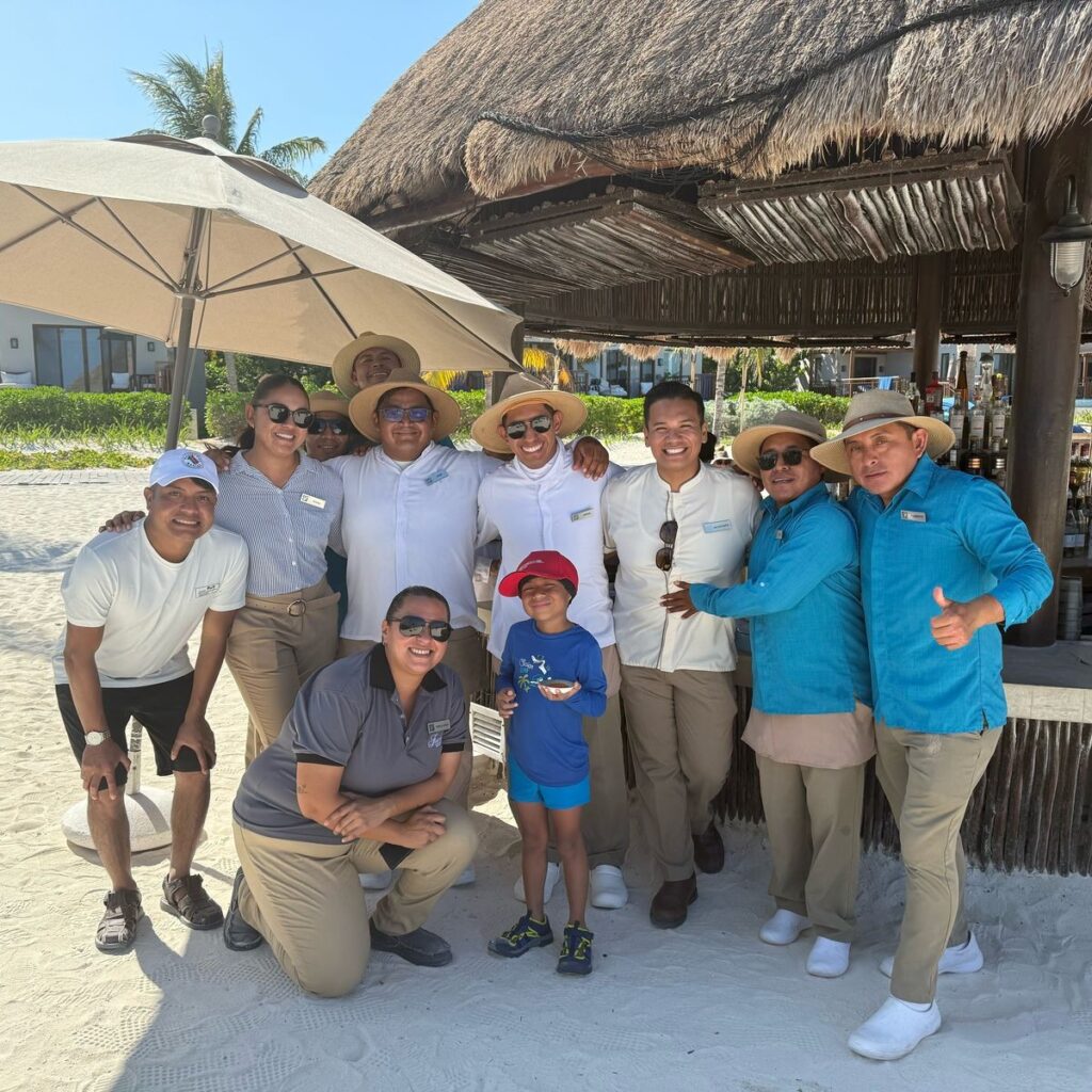 A group of adults and a child pose and smile together on a sandy beach in front of a thatched-roof hut at the Fairmont. Most are wearing uniforms and hats, with palm trees and umbrellas visible in the background.