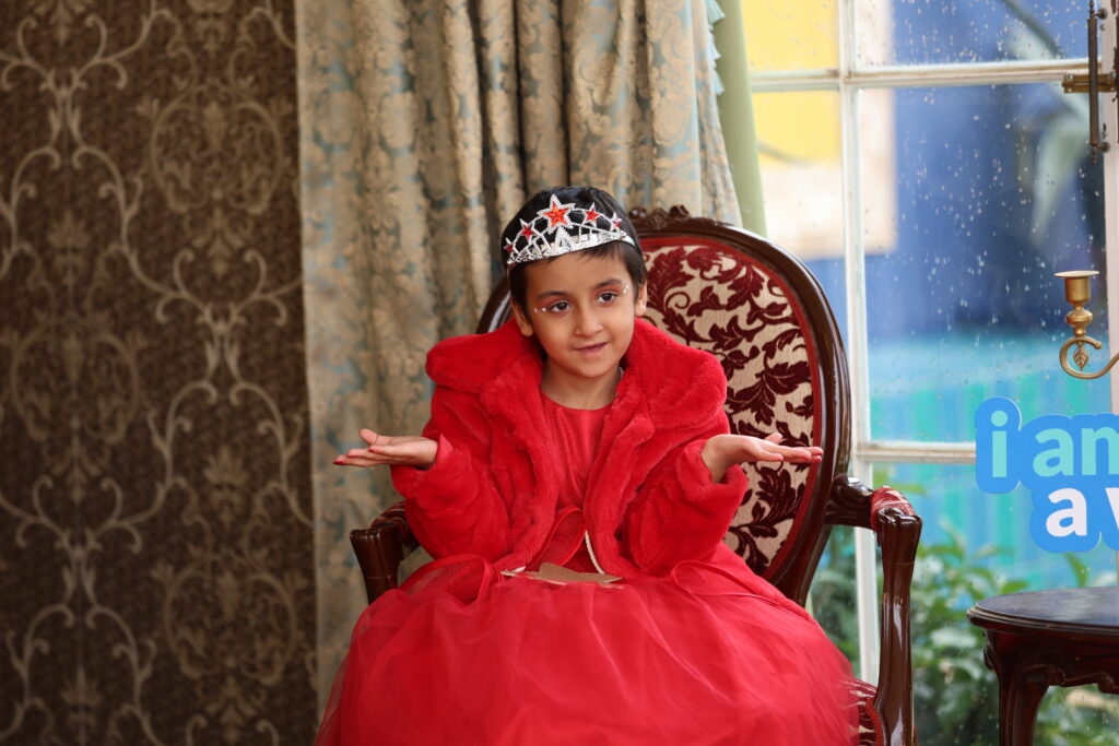 A young girl in a red dress and crown sits on an ornate chair, shrugging with her palms up—perhaps pondering Dimitra's wish to have a new room. She is indoors near a window streaked with raindrops, patterned curtains, and a blue sign in the background.